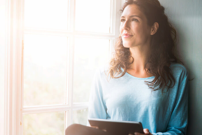 A photo of thoughtful woman sitting on window sill. Young female is holding digital tablet while looking away. She is wearing casuals in brightly lit room.