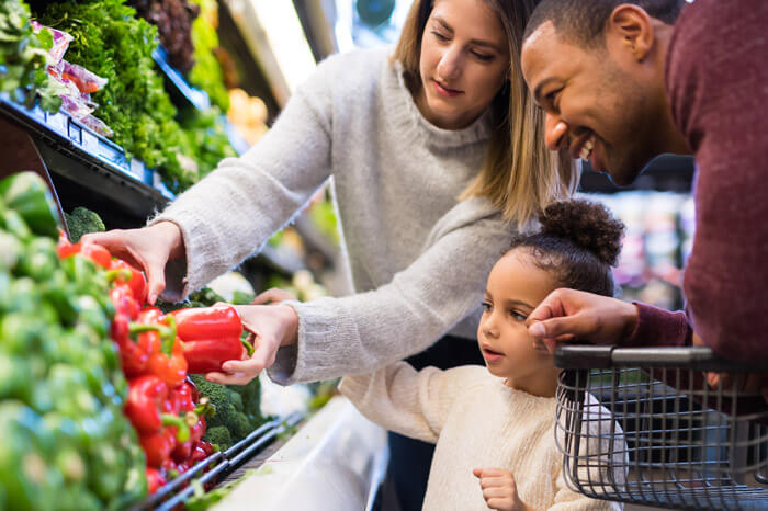 A pre-school age girl helps her parents pick out veggies in the produce section at the grocery store. She is reaching for a red pepper.